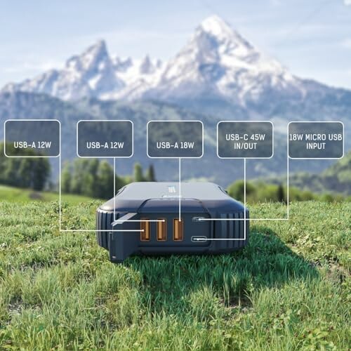 Portable power bank on grass with mountains in background, showing USB ports and inputs.